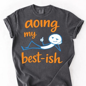 May include: Dark gray t-shirt with the words "doing my best-ish" in orange script. A cartoon figure in white and blue is drawn on the shirt, giving a thumbs up. The shirt has rolled-up sleeves.