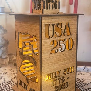 May include: Wooden lantern with cut-out designs and the text "USA 250" and "July 4th 1776-2026". The lantern features a silhouette of the Liberty Bell and the numbers "1776" and "2026".