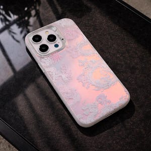 May include: A white phone case with a dragon design. The case has a pearlescent finish with pink and orange tones. The camera cutouts are visible at the top. The case is designed for a smartphone.