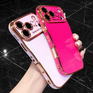 Puede incluir: Dos fundas para teléfono, una rosa y otra blanca, con ribetes dorados. La funda rosa tiene un acabado brillante, mientras que la funda blanca tiene un acabado mate. Ambas fundas tienen el texto "Smile every day" y gráficos de corazón.