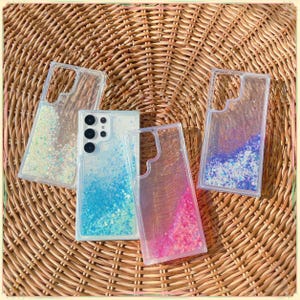 Puede incluir: Fundas transparentes para teléfono con purpurina de colores y un teléfono. Las fundas tienen un efecto de purpurina líquida con purpurina azul, rosa e iridiscente. El teléfono tiene una parte trasera blanca con lentes de cámara negras. Las fundas están sobre un fondo tejido.