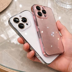 May include: Two smartphone cases, one white and one rose gold, are stacked. The white case has a silver frame and black camera lenses. The rose gold case has a pink frame and black camera lenses. The cases are held in a hand.