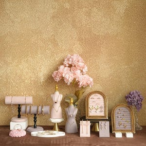 May include: A display of jewelry, including bracelets, necklaces, and earrings, arranged on various stands and busts. The color palette is soft, with pink, gray, and gold accents. A bouquet of pink flowers and a tray of jewelry complete the display.