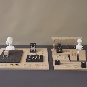 May include: An assortment of jewelry displayed on beige and black platforms. The jewelry includes necklaces, earrings, rings, and bracelets. Some pieces are displayed on small black stands. A white bust is on each side of the display.