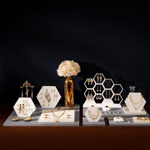 May include: An assortment of jewelry displayed on white and gray hexagonal stands. The jewelry includes necklaces, earrings, bracelets, and rings. A gold vase with white flowers is in the center. The background is a dark navy blue.