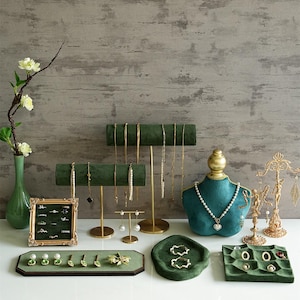 May include: A collection of jewellery display stands and trays in green and gold. The displays showcase necklaces, earrings, and rings. A teal velvet bust holds a pearl necklace. A vase with flowers is on the left. Measurements are in centimetres.