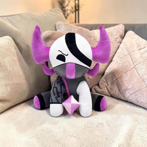 May include: A plush toy with a white head, black eye patch, and purple horns. The toy has a gray body with a geometric pattern on one side, white arms and legs, and a pink gem. The plush is sitting on a beige surface.