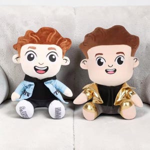 May include: Two plush dolls with cartoon-style faces, brown hair, and smiling expressions. One doll wears a light blue jacket, the other a gold jacket. The dolls have white feet with the words "WHAT'S POPIN" printed on them.