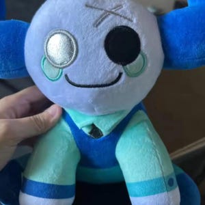 May include: A plush doll with a light blue head, one black eye, and a silver eye. The doll has blue accents on the head and arms, and is wearing a teal and blue outfit. The doll has a stitched mouth and a stitched X on the head.