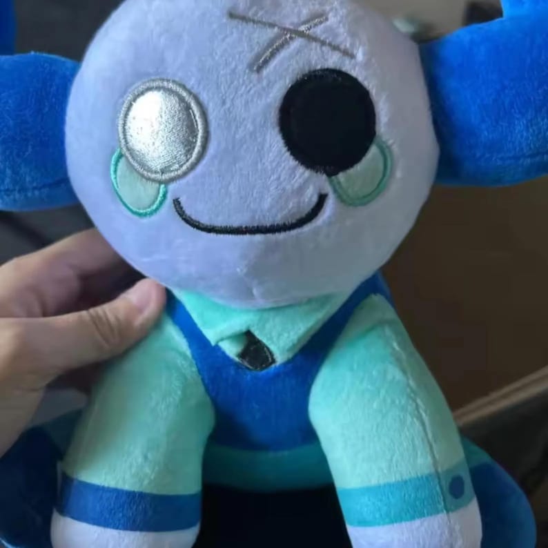 May include: A plush doll with a light blue head, one black eye, and a silver eye. The doll has blue accents on the head and arms, and is wearing a teal and blue outfit. The doll has a stitched mouth and a stitched X on the head.