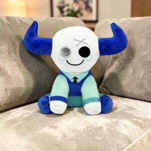 May include: A plush toy in the shape of a bull with a white head, blue horns, and a blue and teal outfit. The bull has a smiling face with one black eye and one gray eye. The toy is sitting on a gray couch.
