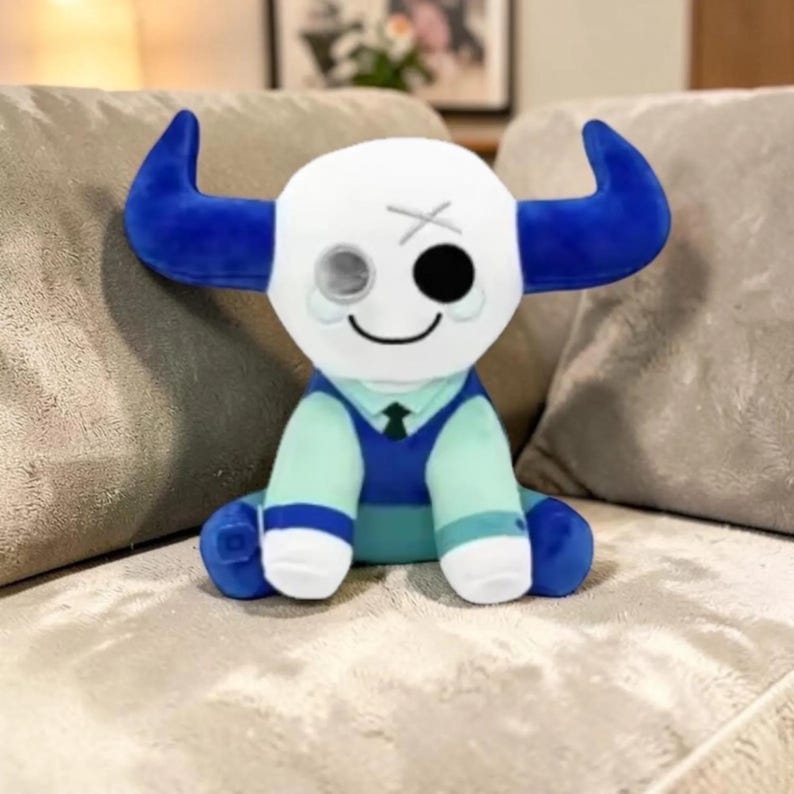 May include: A plush toy in the shape of a bull with a white head, blue horns, and a blue and teal outfit. The bull has a smiling face with one black eye and one gray eye. The toy is sitting on a gray couch.