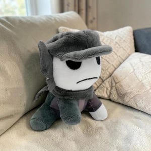 May include: A plush toy character with a white face, black eyes, and a frowning mouth. It wears a gray cap, scarf, and clothing. The plush is sitting on a light-colored couch, with pillows in the background.
