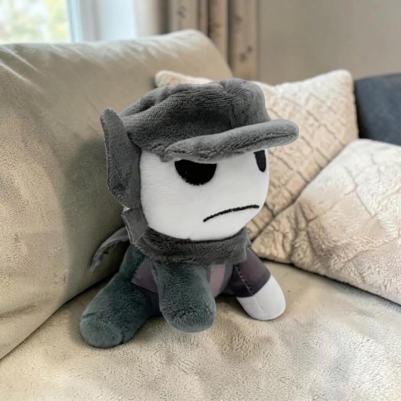 May include: A plush toy character with a white face, black eyes, and a frowning mouth. It wears a gray cap, scarf, and clothing. The plush is sitting on a light-colored couch, with pillows in the background.