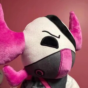 May include: A plush toy with a white head, black accents, and pink horns. The toy has a gray mask and pink details around the mouth and ears. The plushie is soft and cuddly.