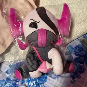 May include: A plush toy in a clear plastic bag. The toy has a white head with black and gray accents, pink ears, and a pink mask. The body is gray with pink details and a geometric pattern on the legs. A pink and white geometric shape is attached.