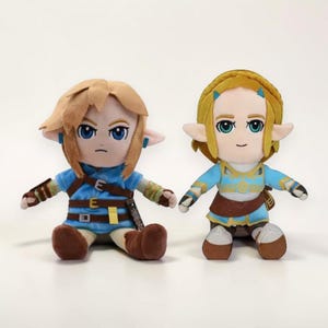 May include: Two plush dolls of video game characters, Link and Zelda, are seated side-by-side. Link has brown hair and a blue tunic with brown accents. Zelda has blonde hair and a blue dress with gold accents. Both dolls have large blue eyes and pointed ears.