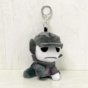 May include: A plush keychain featuring a character with a white face, black eyes, and a stern expression. The character wears a gray hat, scarf, and outfit. The keychain has a silver metal ring for attachment.