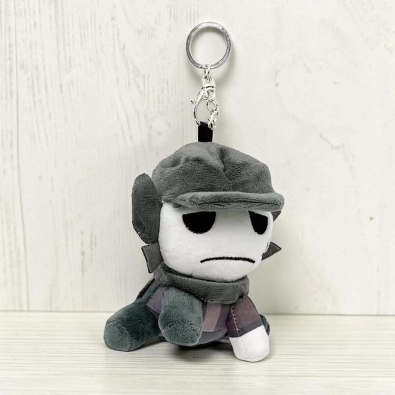 May include: A plush keychain featuring a character with a white face, black eyes, and a stern expression. The character wears a gray hat, scarf, and outfit. The keychain has a silver metal ring for attachment.