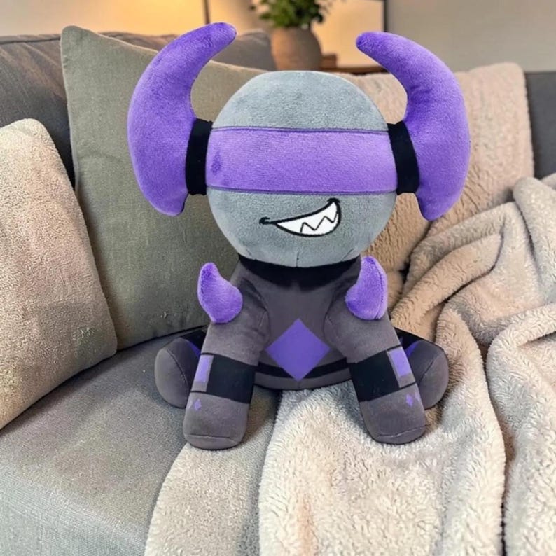 May include: A plush toy in the shape of a cartoon character with a gray head, purple horns, and a purple blindfold. The character has a black and gray body with purple accents and a smiling mouth. The toy is sitting on a gray couch.