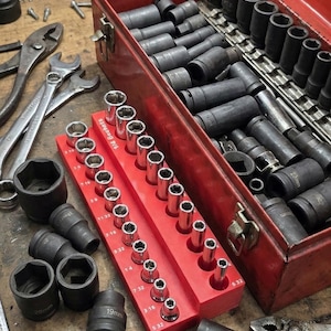 Puede incluir: Una caja de herramientas de metal roja con la tapa abierta, llena de varios vasos y herramientas negros. Un organizador de vasos rojo contiene múltiples vasos. Otras herramientas, incluyendo llaves y alicates, son visibles en la superficie de madera.