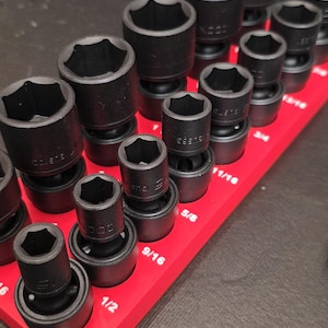 May include: A collection of black impact sockets displayed on a red magnetic holder. The sockets are in various sizes, with measurements in inches visible. A wrench with a black handle and red accents is in the background.