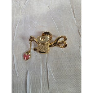 May include: A gold-toned brooch featuring a spool of thread, scissors, and a heart-shaped charm. The brooch has intricate detailing and a pin closure. The heart charm is pink with gold accents. The scissors are open, and the spool is centered.