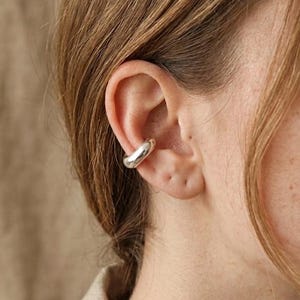 May include: A silver ear cuff is worn on the ear. The ear cuff is a smooth, rounded design. The person's hair is brown, and the background is a neutral color.