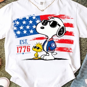 May include: A white t-shirt featuring a graphic of Snoopy and Woodstock wearing sunglasses. Snoopy wears a blue and red varsity jacket. Behind them is a distressed American flag design with the text EST. 1776 in red and blue lettering.