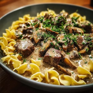 Könnte beinhalten: Nahaufnahme eines herzhaften Gerichts mit Eiernudeln, Rindfleischwürfeln und geschnittenen Pilzen, alles in einer cremigen Sauce. Frische grüne Kräuter sind darüber gestreut, serviert in einer grauen Schüssel.
