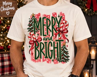 Op de afbeelding: Een crèmekleurig t-shirt met korte mouwen en een grafische print met de tekst MERRY and BRIGHT in groene letters. Het ontwerp bevat twee versierde kerstbomen, twee rood-witte zuurstokken met rode strikken en een rode achtergrond met stippen.