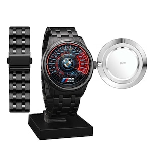 May include: Black wristwatch with a speedometer-style face featuring red and blue accents, the BMW logo, and the letters "M". Includes a separate black watch strap and a silver charging puck with the BMW logo.
