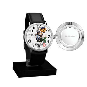 May include: A Polo Ralph Lauren wristwatch with a black leather strap and a white face. The watch face features a bear wearing a golf outfit. The watch is displayed on a black stand and includes a silver-coloured charging component.