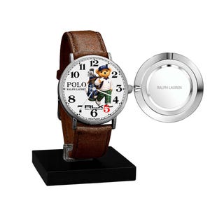 May include: A Polo Ralph Lauren wristwatch with a brown leather strap. The watch face features a bear wearing a golf outfit. The watch is displayed on a black stand. The watch face has the numbers 1-12 and the words "POLO RALPH LAUREN".