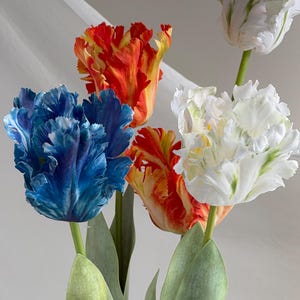 May include: A bouquet of colorful tulips with ruffled petals. The flowers display vibrant hues of blue, orange, and white, with green stems and leaves. The tulips are arranged against a soft, neutral background, creating a visually appealing floral arrangement.