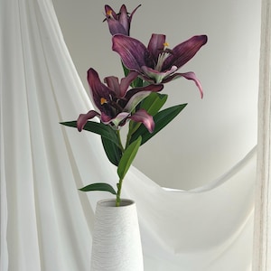 May include: A white textured vase holds a stem of artificial lilies. The lilies have deep purple petals with yellow stamens and green leaves. The vase is on a white surface with a white curtain in the background.