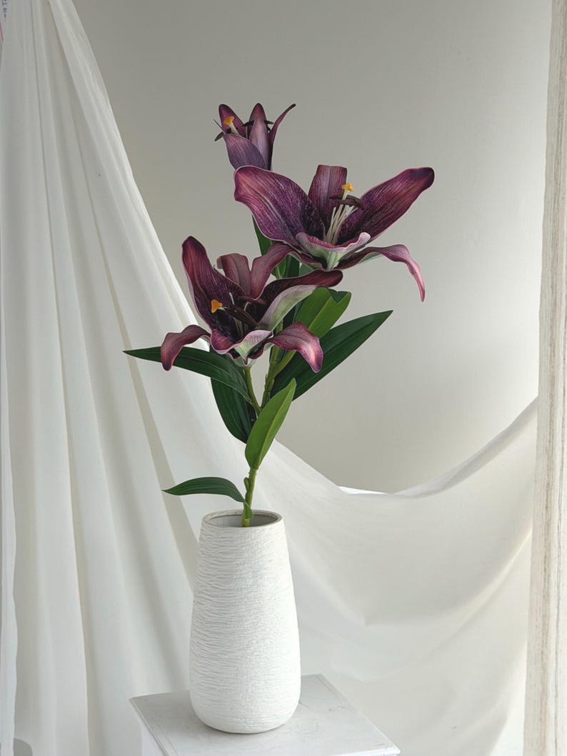 May include: A white textured vase holds a stem of artificial lilies. The lilies have deep purple petals with yellow stamens and green leaves. The vase is on a white surface with a white curtain in the background.