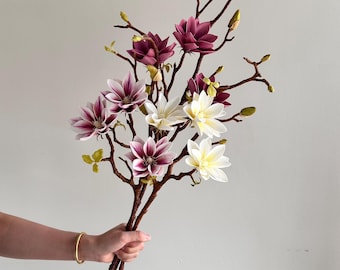 May include: A decorative branch arrangement featuring multiple magnolia flowers in shades of deep magenta, light purple, and creamy white. The stems are brown and textured, accented with small green buds throughout the branching structure.
