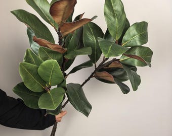 May include: A branch of artificial magnolia leaves featuring large, glossy, dark green foliage on the top side and a contrasting matte, brownish-copper underside. The stems are thin and brown, supporting multiple clusters of broad, oval-shaped leaves.