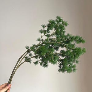 May include: A sprig of artificial greenery with multiple branches and small, needle-like leaves. The leaves are a vibrant green, and the branches are a muted brown. The sprig is held against a neutral background, suggesting a decorative or craft item.