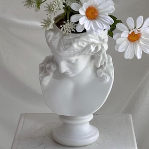 May include: A white bust vase in the shape of a classical woman's head and shoulders, holding white and yellow daisies and other white flowers. The vase has a pedestal base and is placed on a white surface.