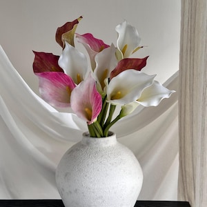 May include: A bouquet of artificial calla lilies in shades of white, pink, and deep maroon, each featuring a central yellow spadix. The flowers are arranged in a round, textured, off-white ceramic vase with a narrow neck and a spherical body.