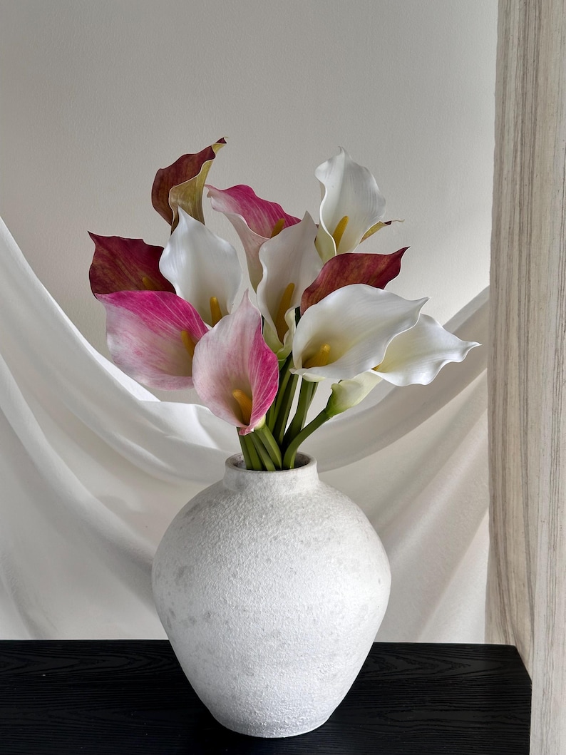 May include: A bouquet of artificial calla lilies in shades of white, pink, and deep maroon, each featuring a central yellow spadix. The flowers are arranged in a round, textured, off-white ceramic vase with a narrow neck and a spherical body.