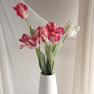 May include: A white textured vase holds a bouquet of tulips. The tulips have ruffled petals in shades of pink, red, and white, with green stems and leaves. The vase is on a white surface, with a white curtain in the background.