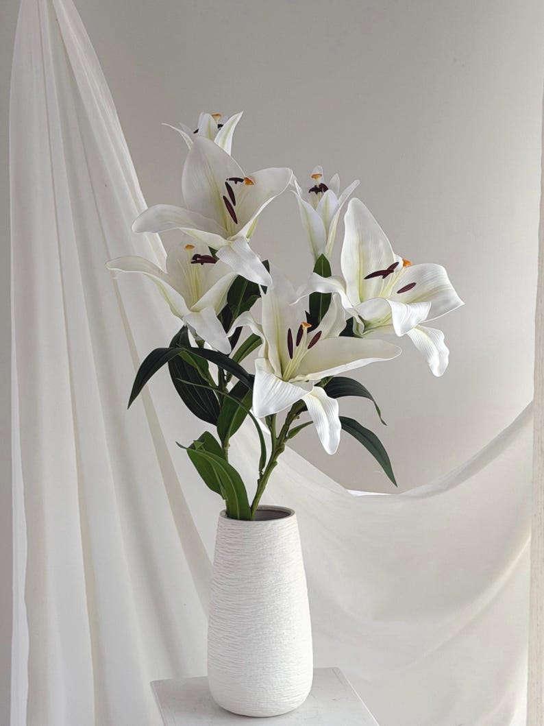 May include: A bouquet of white lilies with dark green leaves in a textured white vase. The lilies have prominent stamens and are arranged against a backdrop of white fabric, creating a soft, elegant aesthetic.