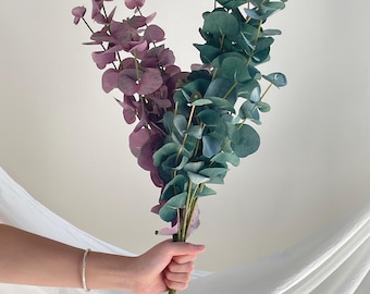 Op de afbeelding: Een bundel kunstmatige eucalyptusstengels met twee verschillende kleurvariaties. De ene set stengels heeft gedempte paarse, ronde bladeren, terwijl de andere set diepgroene, blauwgroene, ronde bladeren op slanke, rechtopstaande stelen heeft.