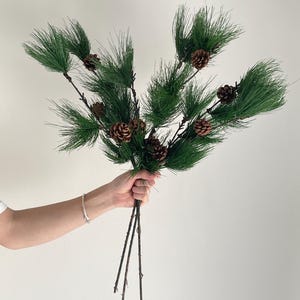 May include: A bundle of artificial pine branches featuring long, thin, dark green needles and several small, brown, textured pinecones attached to the stems. The branches are arranged in a fan-like shape with thin, brown, woody-looking stalks.