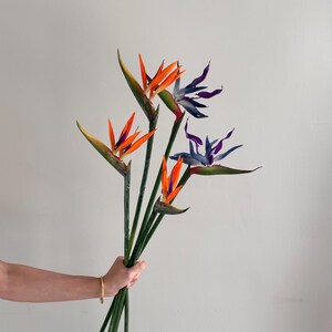 Op de afbeelding: Een boeket kunstmatige paradijsvogelbloemen met drie oranje en blauwe bloemen en twee paarse en blauwe bloemen, bevestigd aan lange groene stelen. De bloemen hebben puntige, bloembladachtige structuren en groene schutbladeren.