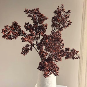 Peut inclure: Un vase blanc texturé contient un arrangement décoratif de branches avec de petites baies brun-rouge. Le vase a une forme arrondie et une texture verticale rugueuse. L'arrangement est placé sur un fond neutre.