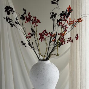 May include: A white, textured ceramic vase contains branches with red berries and dark leaves. The vase is on a white pedestal, with a white curtain behind. The branches have a natural, organic appearance.